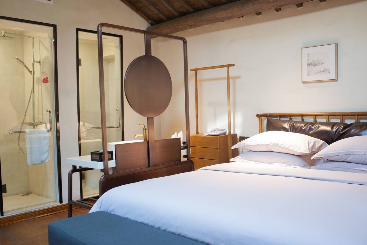 Una camera d'albergo con un letto, un comodino, uno specchio, una testiera in legno, un'area doccia.