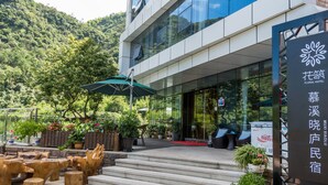 A hotel entrance with a modern glass facade, steps leading up to the entrance, outdoor seating area with wooden furniture, and potted plants. - Floral Hotel Hangzhou Muxixiaolu (Hangzhou)