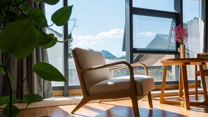A room with a wooden floor, a beige armchair, a wooden table, and a large window with a view of buildings and hills. - Floral Hotel Hangzhou Muxixiaolu (Hangzhou)