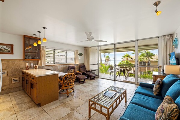 A spacious living area with a large sliding glass door leading to a balcony, a blue sofa, a wooden coffee table, and a kitchen area with a dining table and chairs. - Across From Kamaole 2 Beach, This Concrete Resort Is Maintained Well (Kihei, Maui)
