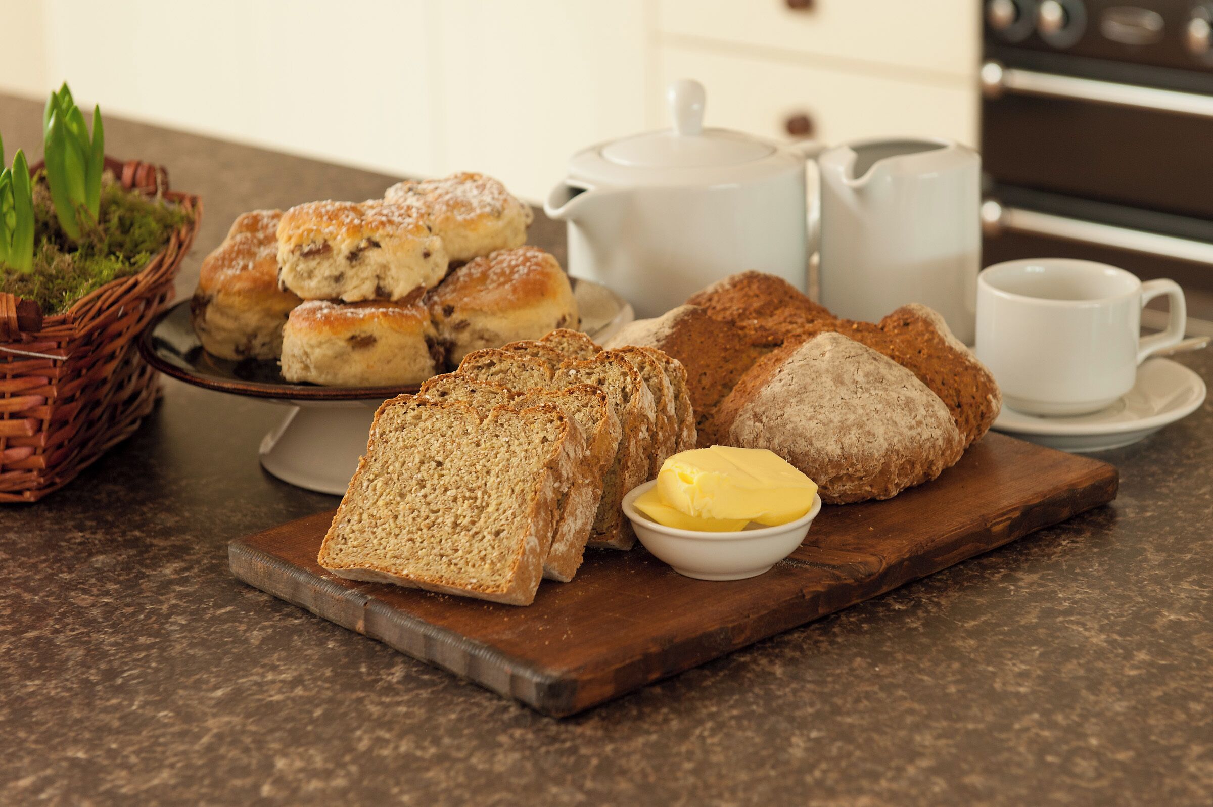Une table dressée pour le petit-déjeuner, avec du pain, du beurre et des viennoiseries disposés sur une planche en bois, accompagnés d’une théière et de tasses.