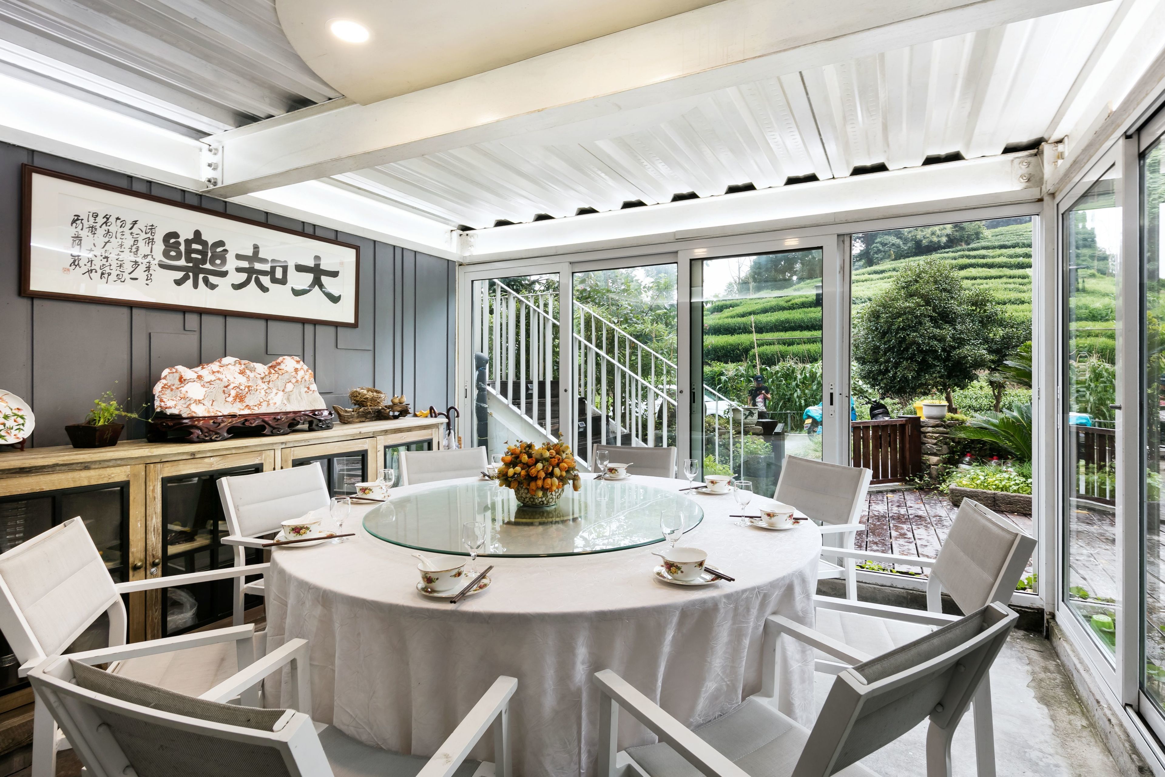 A round dining table with white chairs, a glass top, and a vase of flowers. There is a staircase leading to a garden with a view of green hills.