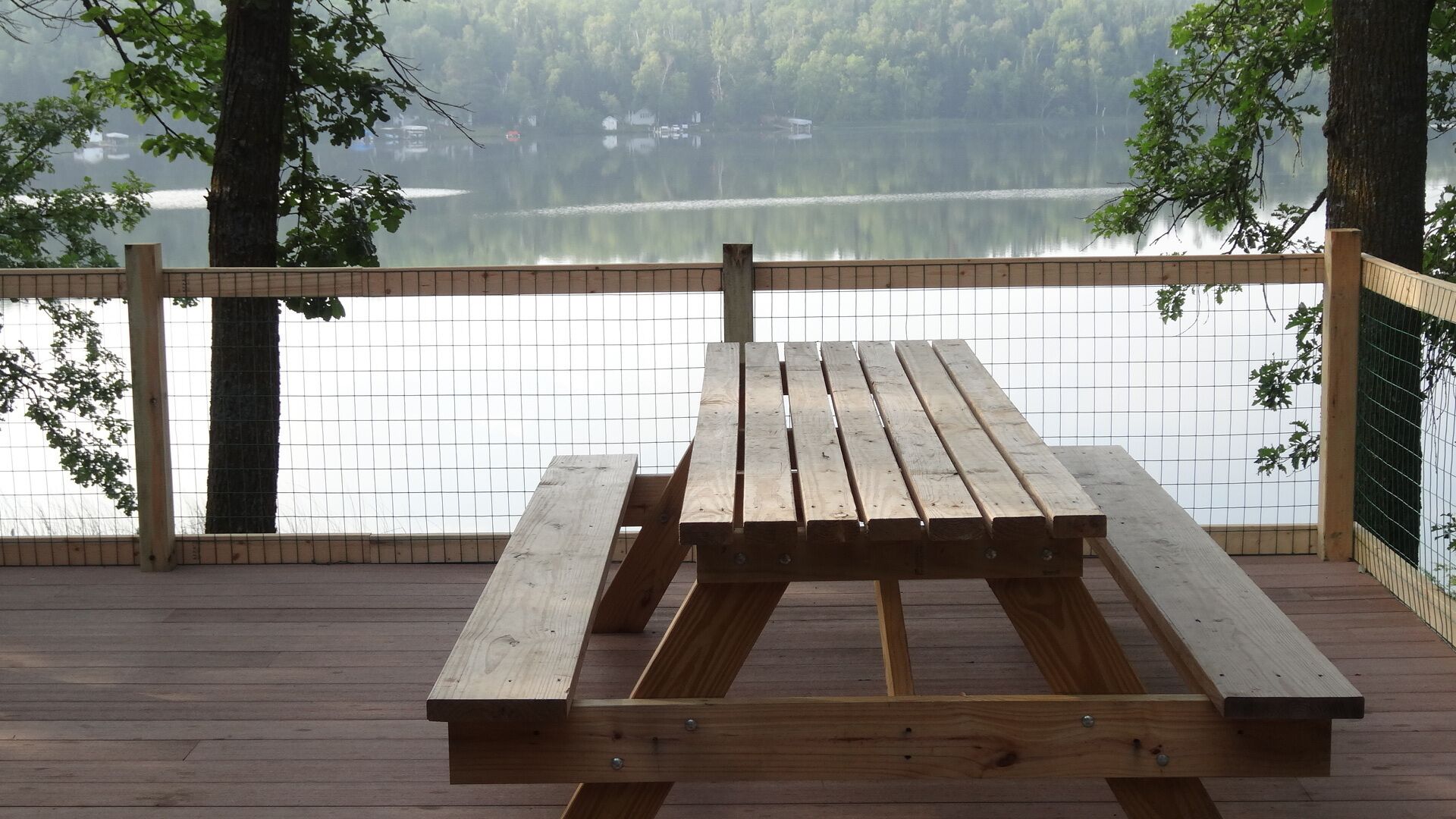 Una mesa de picnic de madera en un muelle con vista a un lago tranquilo, una cerca y árboles.