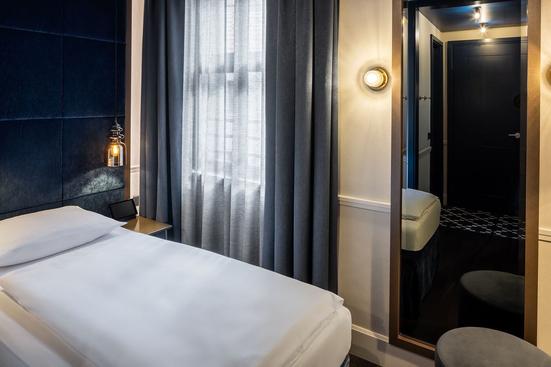 A hotel room with a bed, a nightstand with a lamp, a bathroom with a toilet and sink, and a checkered floor.