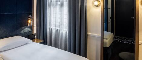 A hotel room with a bed, a nightstand with a lamp, a bathroom with a toilet and sink, and a checkered floor.