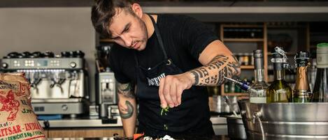 Una persona preparando comida en una cocina con estufa, cafetera y varias botellas.