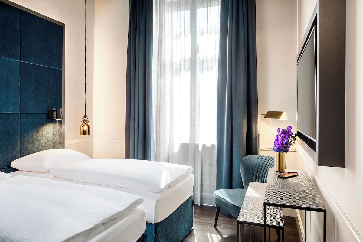 Habitación de hotel con dos camas, un escritorio, una silla, una lámpara y un ventanal grande con cortinas.