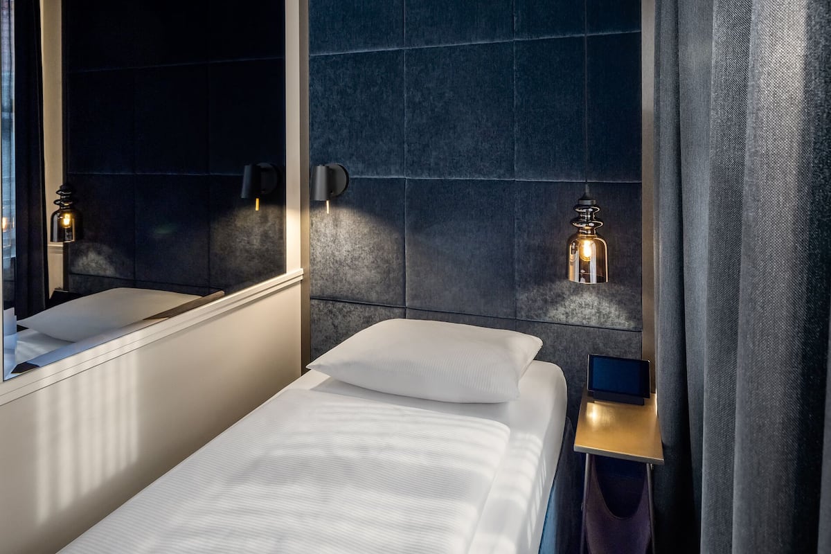 Una habitación de hotel moderna con un espejo grande, una mesita de noche con una lámpara, y otra mesita de noche con una pantalla.