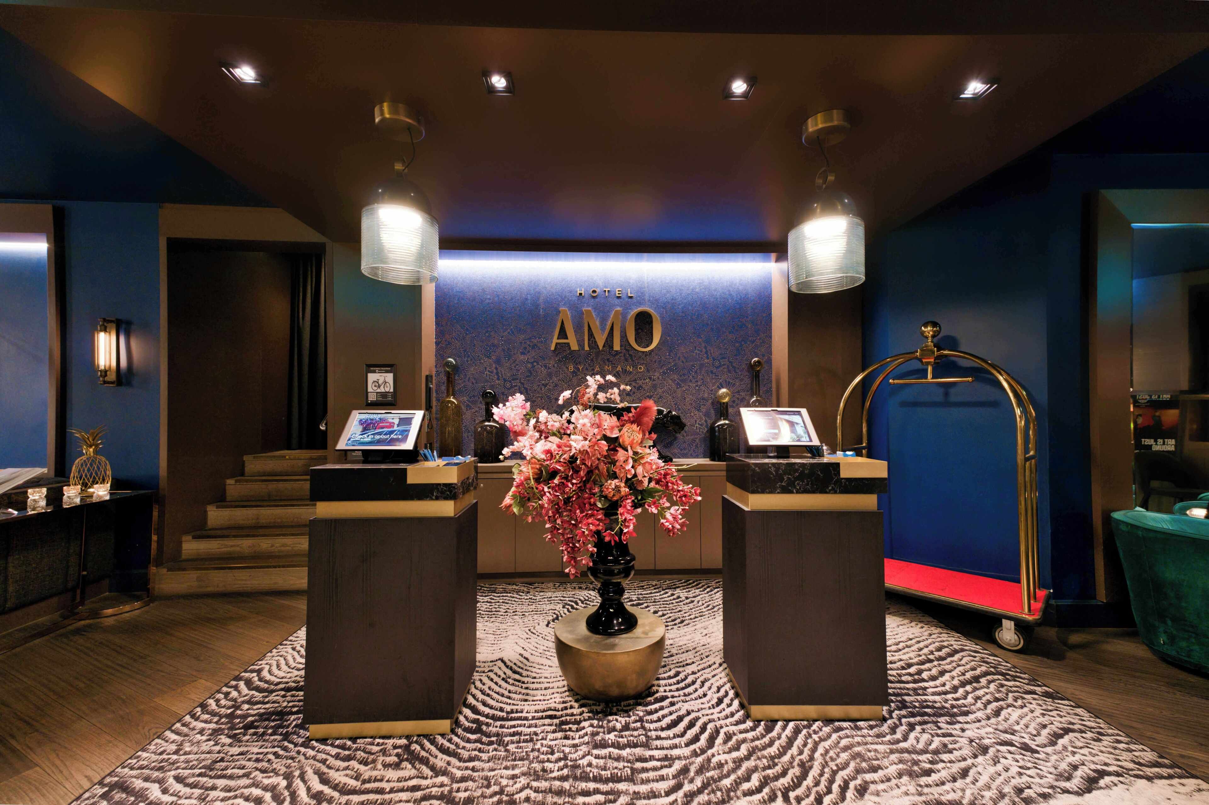 A hotel reception area with a reception desk, a digital screen, a flower arrangement, and a cart.