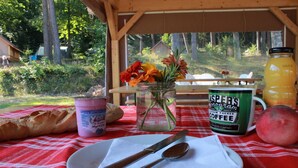 A picnic setup with a jar of flowers, a can of coffee, a bottle of juice, a baguette, and a plate with cutlery on a red and white checkered tablecloth.