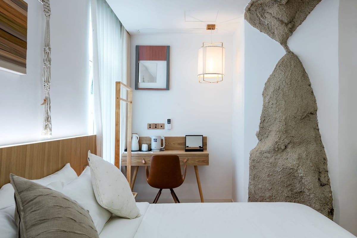 Une chambre d’hôtel comprenant un lit, un bureau, une chaise, une décoration murale en macramé, un tableau encadré et une sculpture de sable originale.