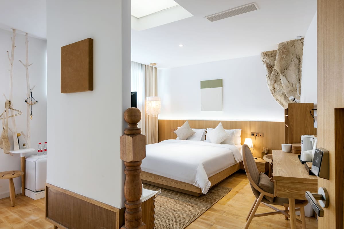 Une chambre d’hôtel moderne avec un lit en bois, un bureau avec une chaise, une petite table et une grande fenêtre.