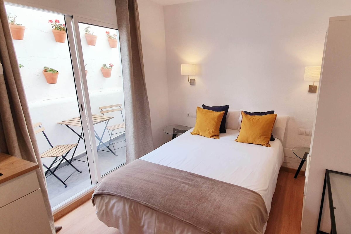Un dormitorio con una cama, dos lámparas de noche, un escritorio, una silla y un ventanal corredero que da a un espacio exterior con plantas en macetas.