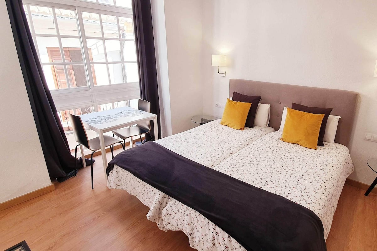 Un dormitorio con cama, lámparas de noche, una mesita y una ventana con cortinas.
