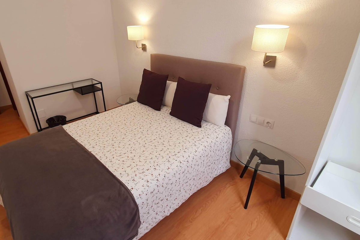 Una cama bien tendida con cubrecama floral, dos lámparas de noche, una mesita y suelo de madera.