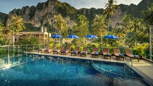 A swimming pool with lounge chairs and umbrellas, surrounded by palm trees and a mountainous backdrop. - Andaman Breeze Resort - (Krabi)
