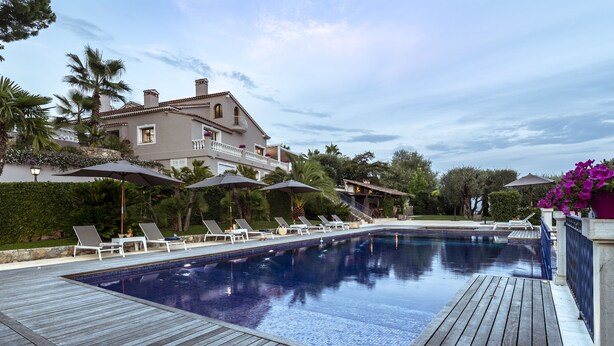 Una piscina con sillas de descanso y sombrillas, rodeada por una terraza de madera y una casa grande con balcón.