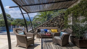 Un área de estar al aire libre con techo, mobiliario de mimbre y un muro de piedra como fondo.