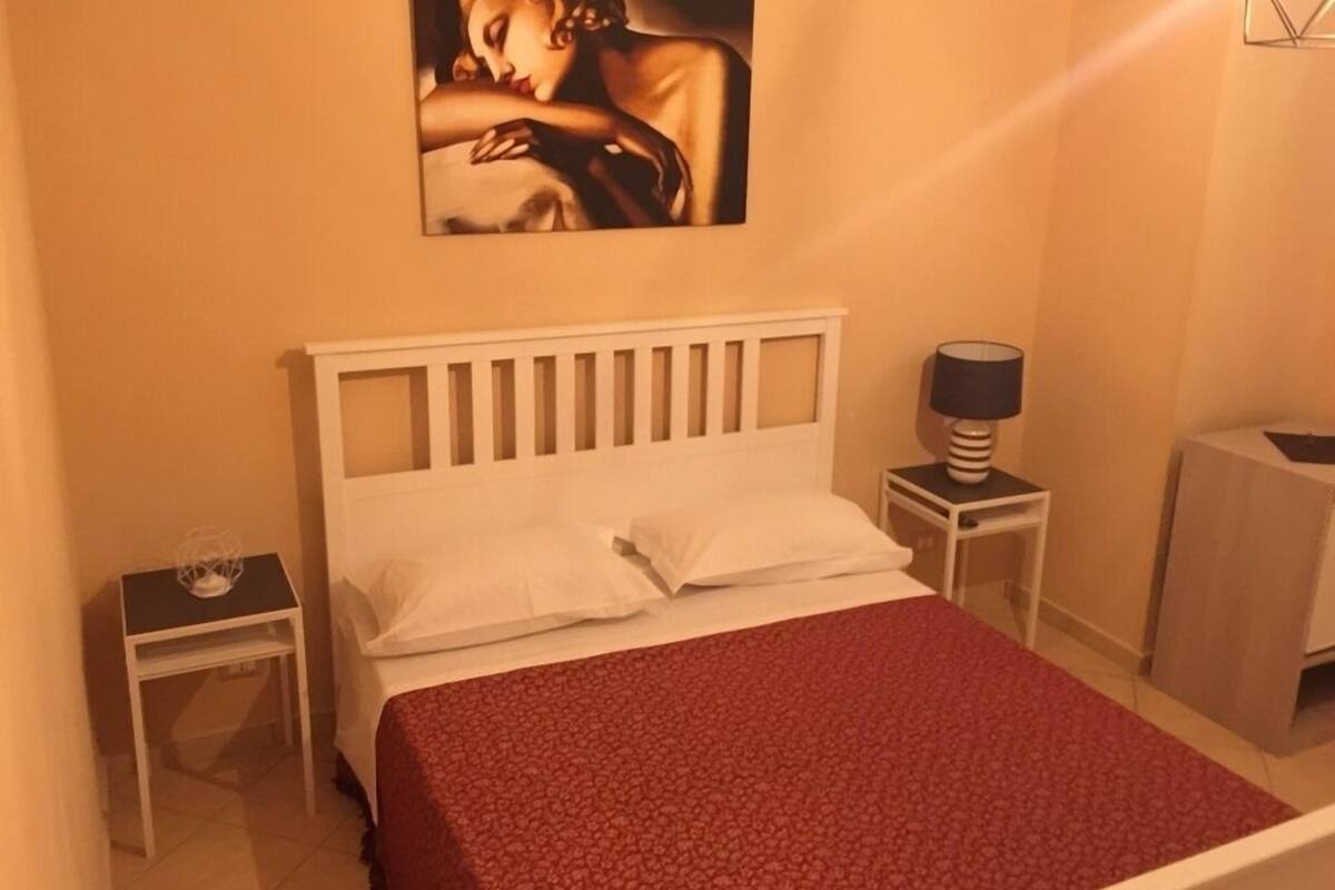 Un dormitori amb un llit blanc, un llençol vermell estampat, un quadre a la paret, una nitòria amb una làmpada i una taula petita amb un objecte decoratiu.