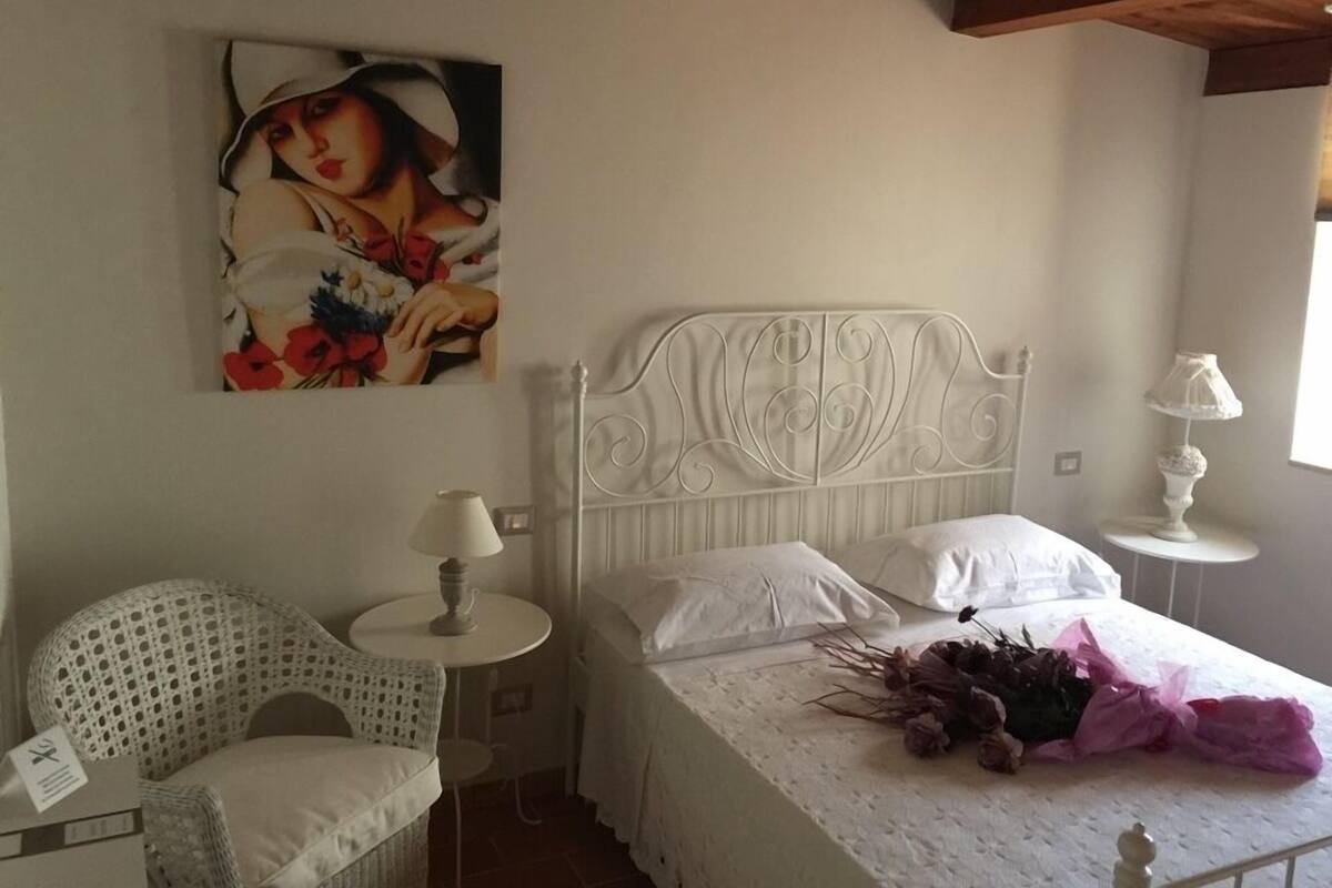 Un dormitori amb llit blanc, capçalera blanca, taula rodona petita amb una làmpada, cadira de vimet i un quadre a la paret.