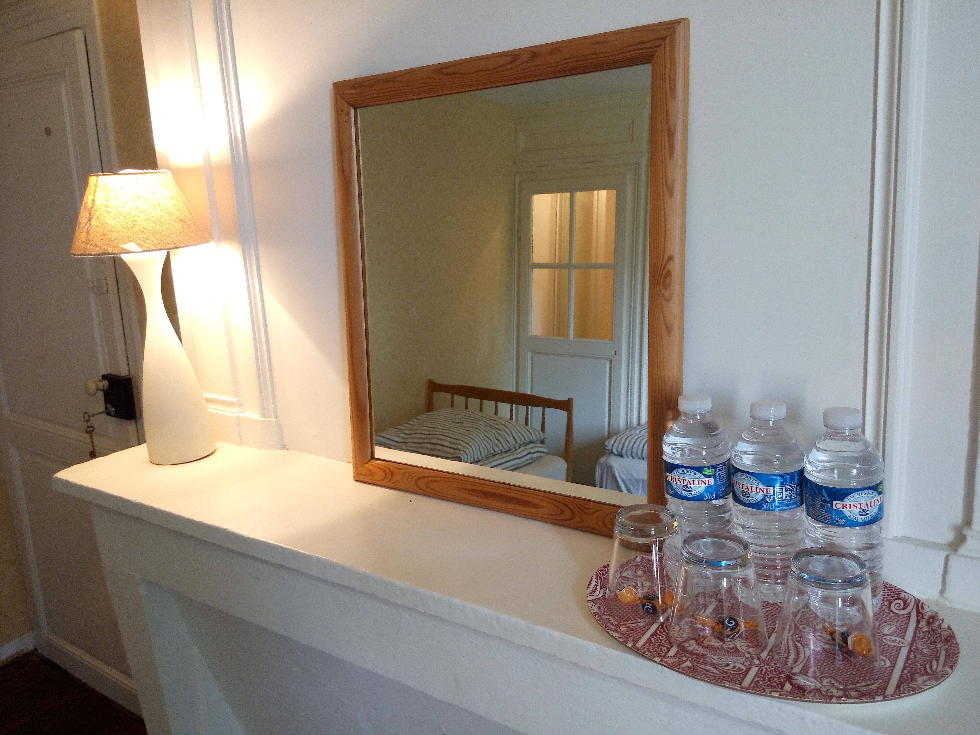 Une chambre avec un miroir à cadre en bois, une table de chevet blanche, une lampe et un plateau contenant des bouteilles d’eau et des objets décoratifs.