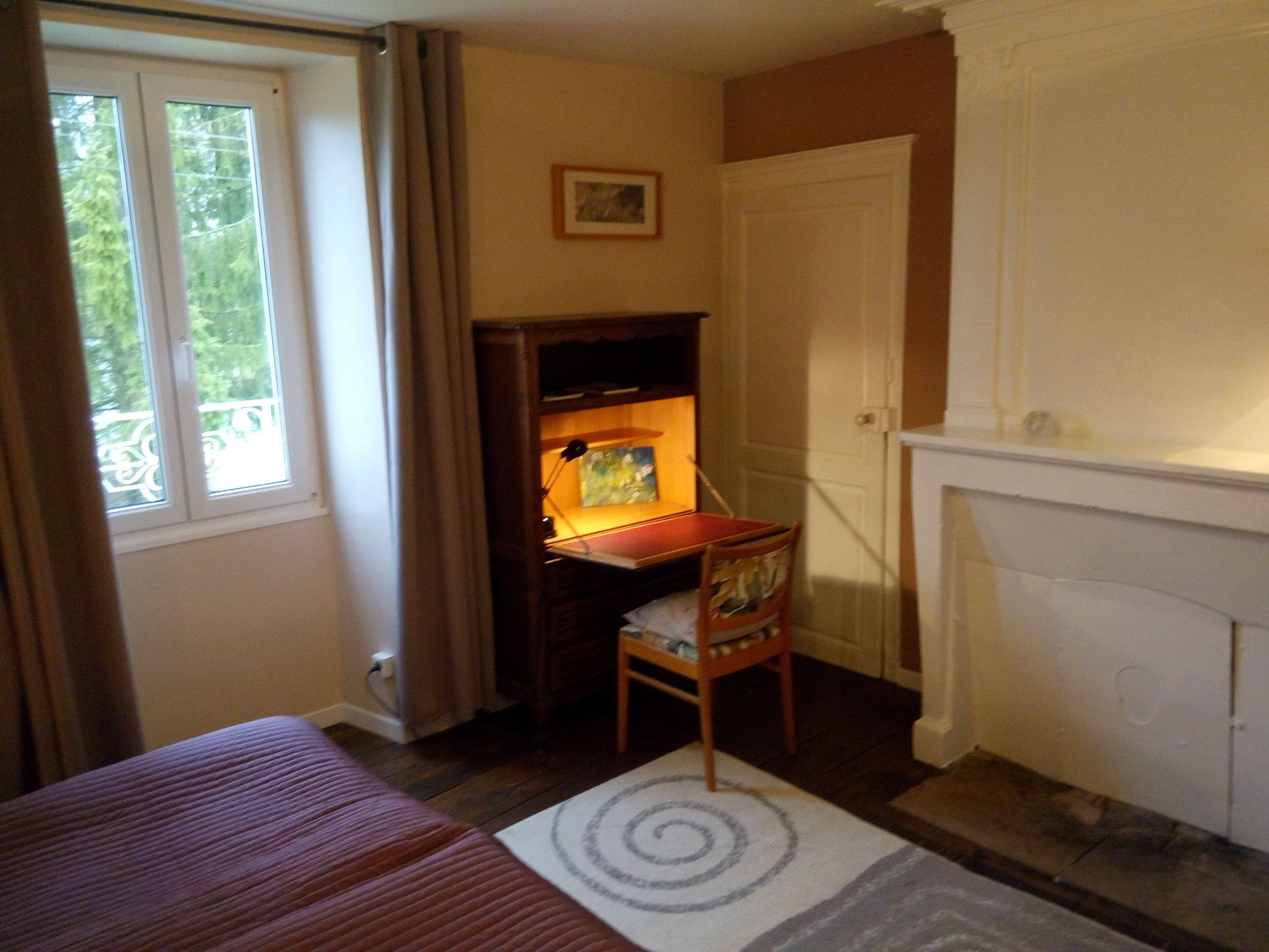 Una habitación con cama, escritorio con lámpara, una silla, chimenea y ventana con cortinas.