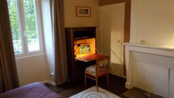 Una habitación con cama, escritorio con lámpara, una silla, chimenea y ventana con cortinas.