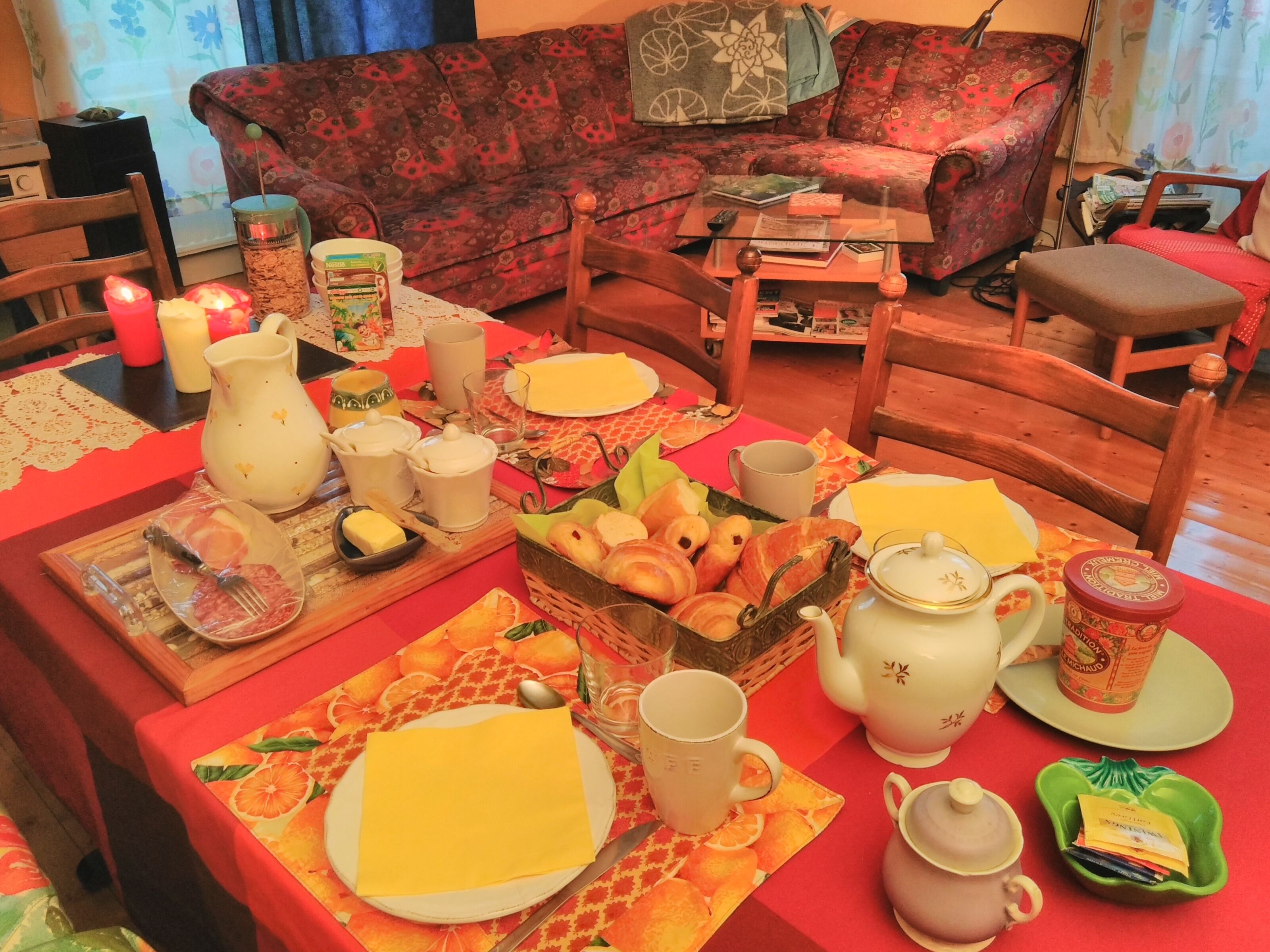 A dining table set with plates, cutlery, and a variety of food items, surrounded by wooden chairs and a sofa in the background.