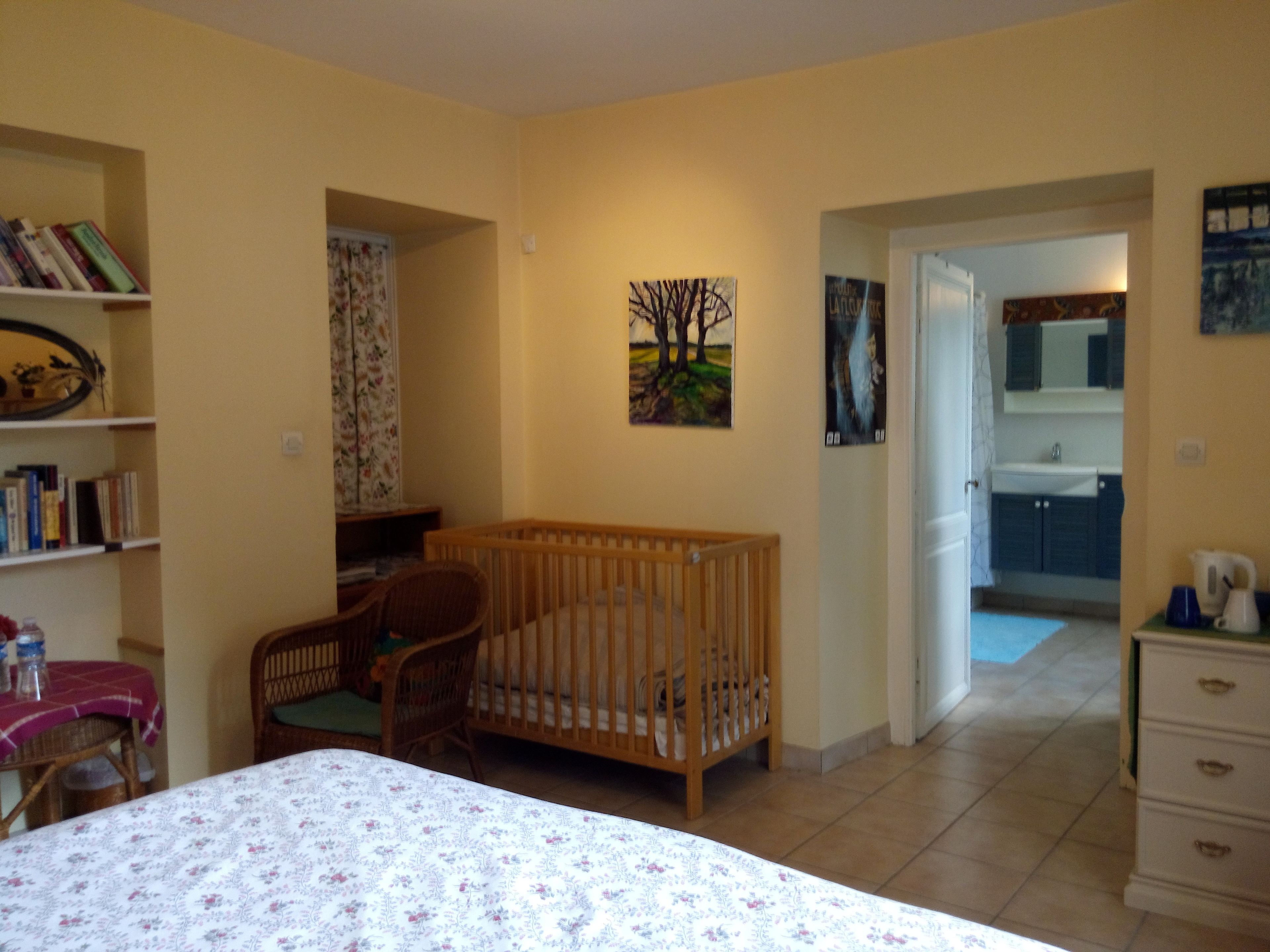 Ein Zimmer mit einem Babybett, einem Sessel, einem Tisch mit Blumenmuster und einem Bücherregal.