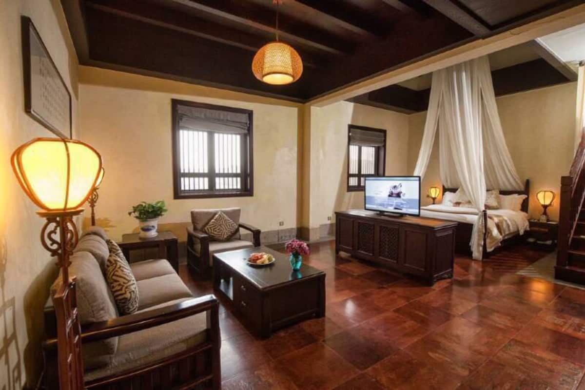 Ein geräumiges Wohnzimmer mit einer Couch, einem Couchtisch und einem Fernseher. Im Hintergrund ist ein Bett mit Baldachin zu sehen.