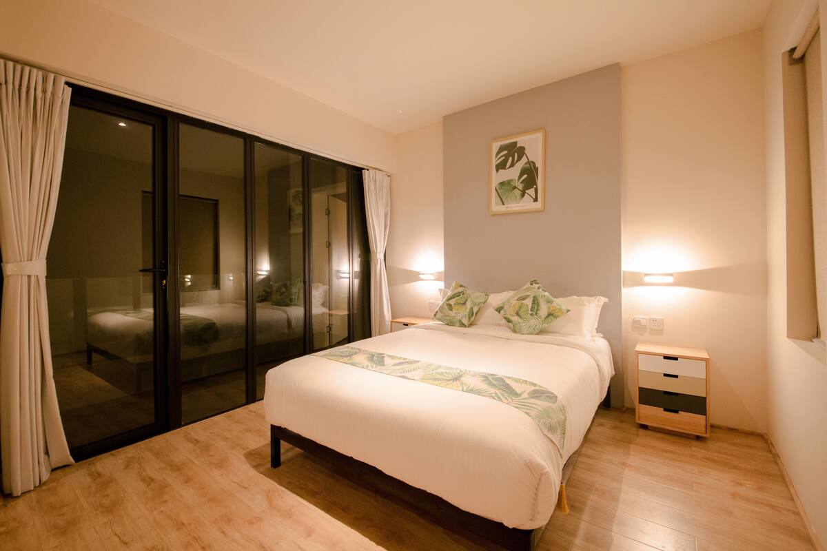 A bedroom with a large bed, a sliding glass door leading to a balcony, a bedside table with a lamp, and a wall-mounted artwork.
