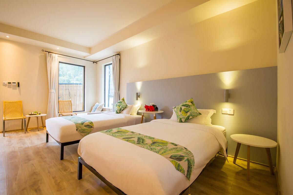 A hotel room with two beds, a small table, a chair, and a balcony with curtains.
