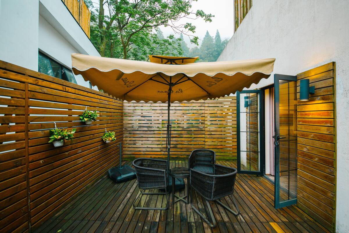 A wooden deck with a table and chairs under a large umbrella, surrounded by wooden slats and potted plants.