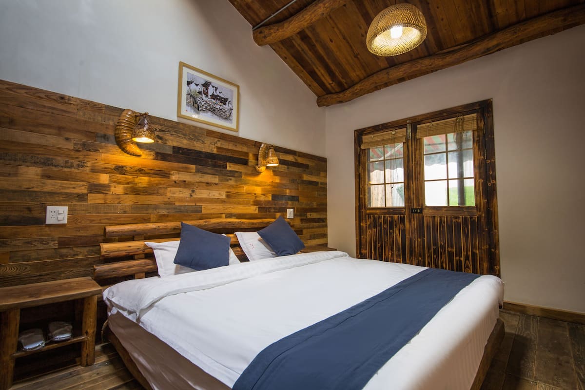 Ein Bett mit Holzrahmen, weißer Bettwäsche und blauen Akzenten, ein Holzkopfbrett mit wandmontierten Lampen, ein kleiner Nachttisch und ein Fenster mit Holzjalousien.