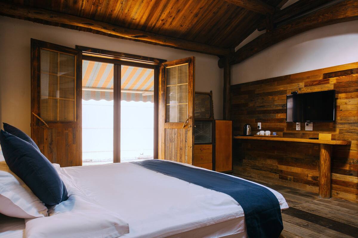 Ein Schlafzimmer mit Holzbalkendecke, einem Bett mit weißer Bettwäsche, einem hölzernen Schminktisch und einem Fernseher an der Wand.