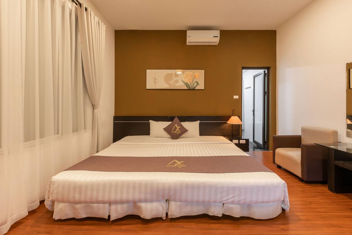 A hotel room with a large bed, a desk, and a chair. There is a painting on the wall and an air conditioning unit above the bed.