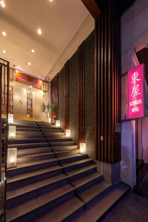 A hotel entrance with a staircase leading to the lobby, a neon sign, and a stone wall.