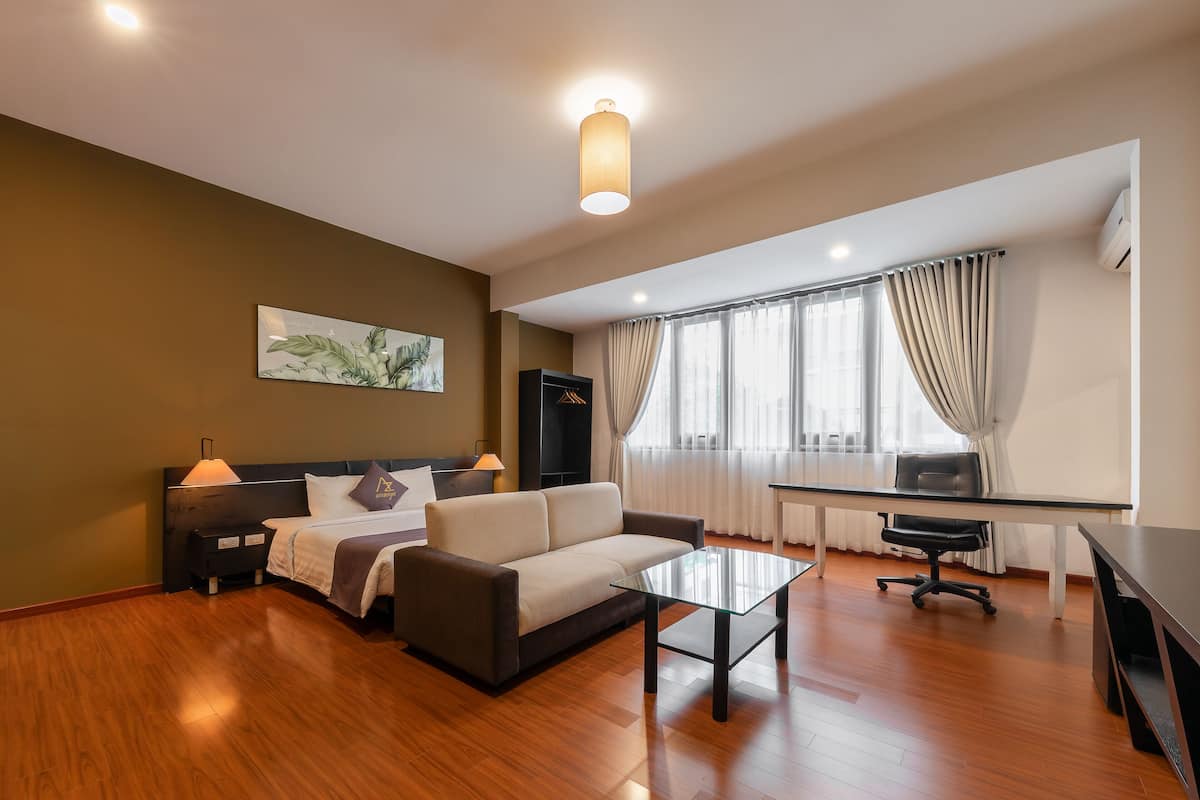 A modern hotel room with a sofa, a glass coffee table, a desk, and a large window with curtains.