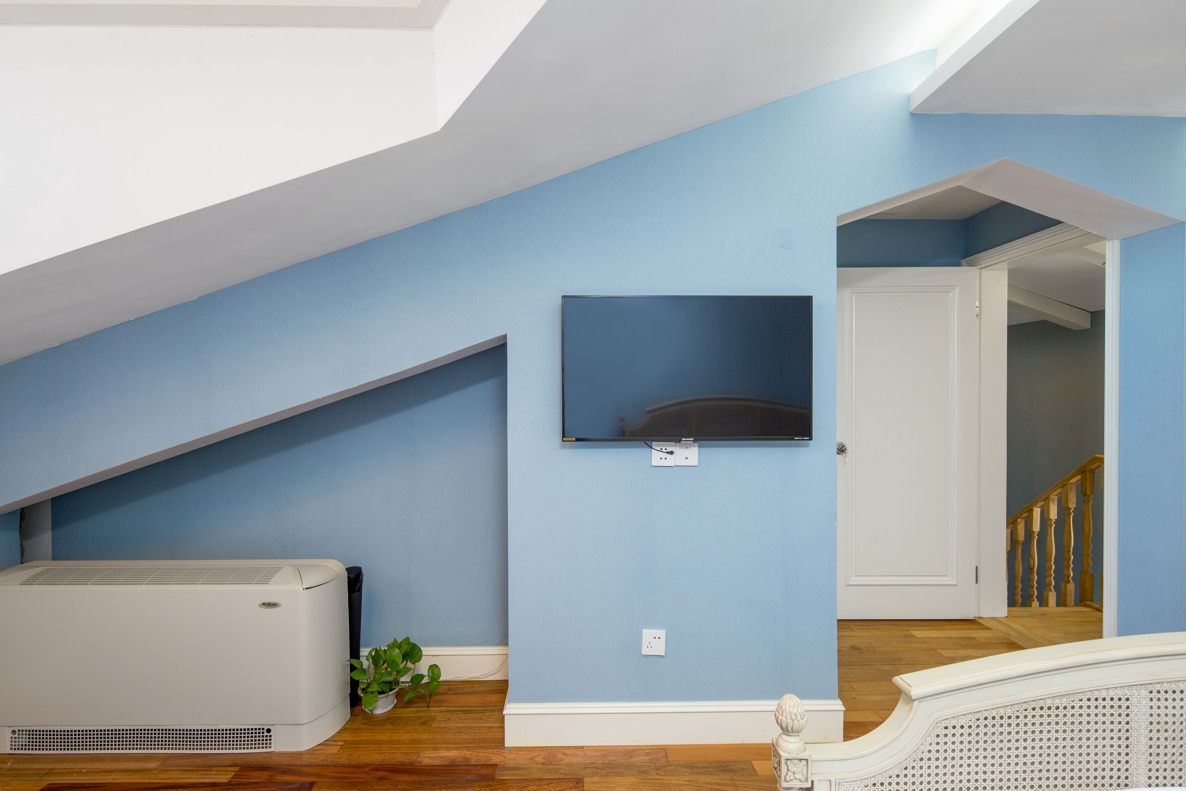 Una habitación con techo inclinado, un televisor de pantalla plana montado, un aire acondicionado blanco, suelo de madera y una puerta blanca que da a otra habitación.