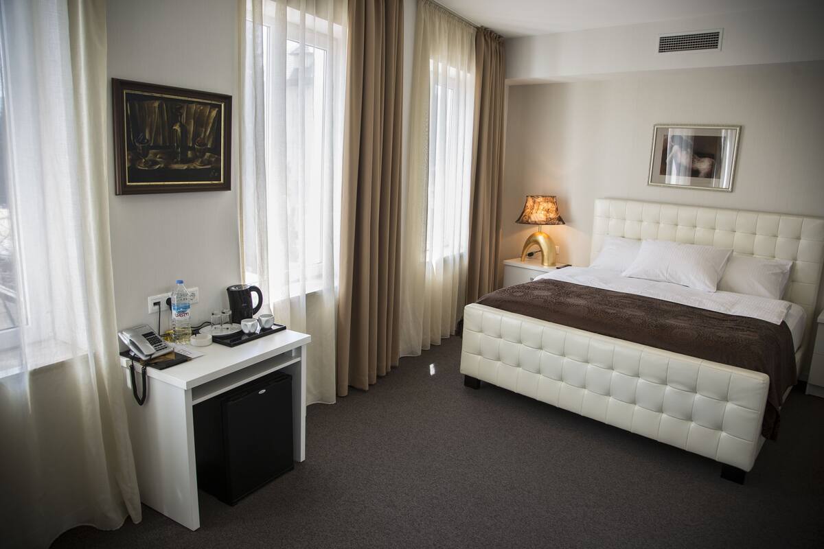 A hotel room with a white bed, a desk with a coffee maker, a lamp, and a painting on the wall.