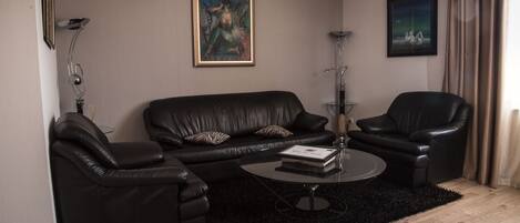 A modern living room with a black leather sofa, two black leather armchairs, a round coffee table, and a black rug. There are two framed artworks on the walls.