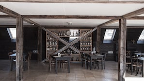 Une pièce spacieuse avec un plafond haut, des poutres en bois et un escalier central menant à un niveau supérieur. Des tables et des chaises y sont disposées pour les repas ou les moments de convivialité.