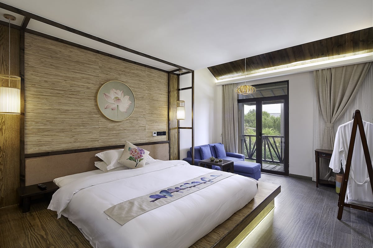 Ein Hotelzimmer mit einem großen Bett, einem blauen Sessel, einem Holzkopfschild, einem Nachttisch und Blick ins Freie durch ein großes Fenster.