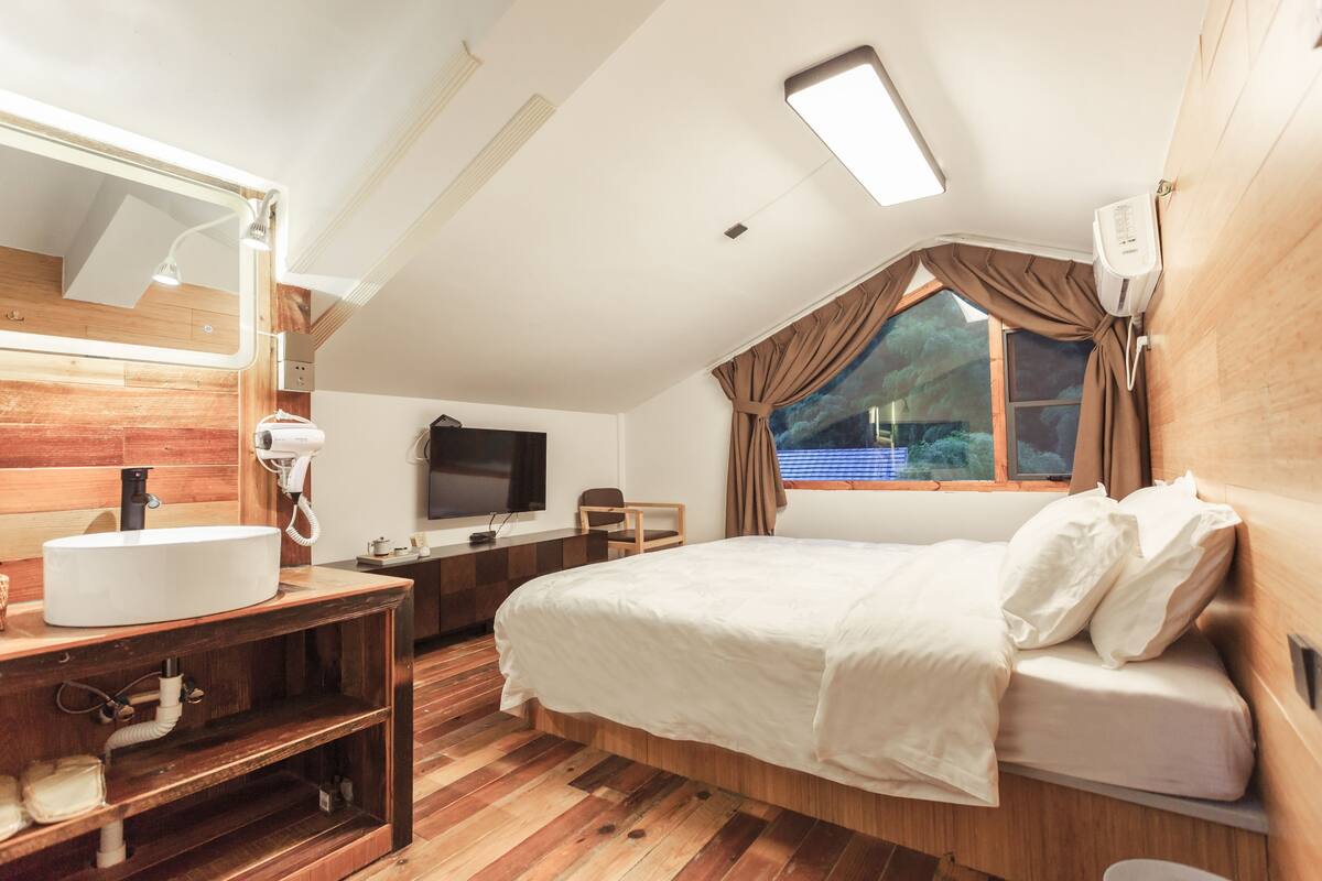 A bedroom with a wooden floor, a bed with white bedding, a bedside table, a mirror, a TV, and a window with curtains.