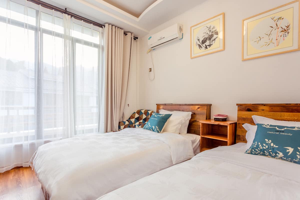 A hotel room with two beds, a wooden headboard, a bedside table with a lamp, and a window with sheer curtains.