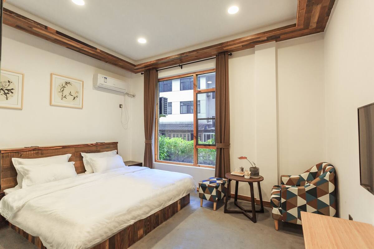 A bedroom with a large bed, a window with curtains, a small table with a plant, and a patterned armchair.