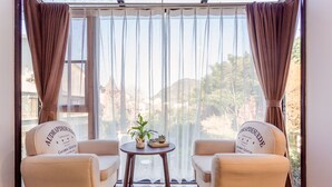A room with two beige armchairs facing a large window with sheer curtains, a small round wooden table with a plant and a tray, and a view of a forested area. - That Mountain That Shed (Huzhou)