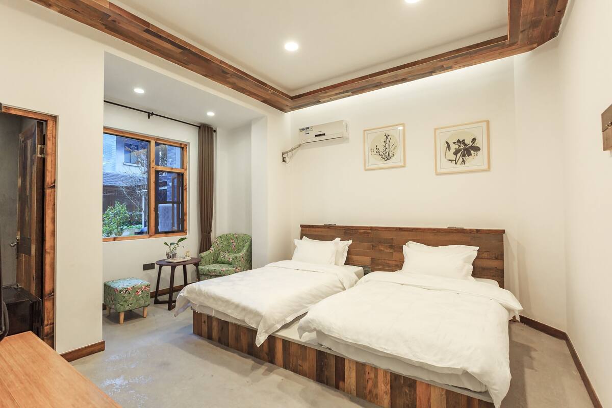A bedroom with two beds, a wooden ceiling, an air conditioning unit, and a window with a view of greenery.