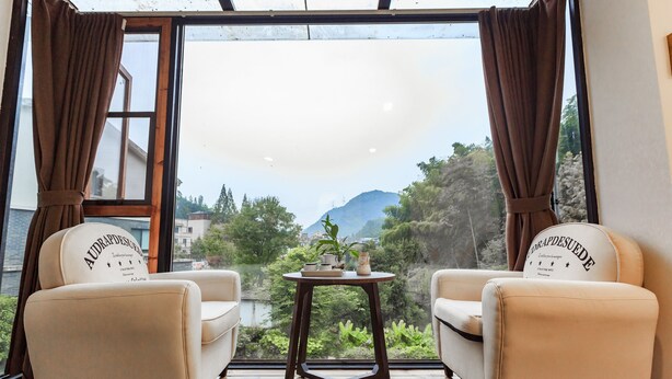 A room with two beige armchairs facing a large window with curtains, a small round table with a plant, and a view of a mountainous landscape. - That Mountain That Shed (Huzhou)