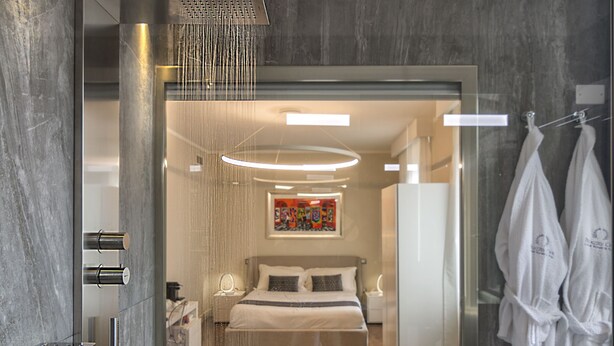 A modern bathroom with a glass-enclosed shower, a bed, a seating area, and a robe rack. - Palazzo Cini Luxury Rooms In Pisa (Pisa)